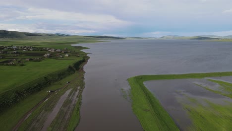 Aerial-shot-of-a-lake-surrounded-by-meadows-villages-old-houses-mountains-river