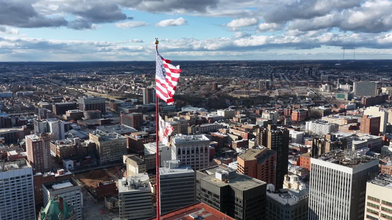 Vista aerea di una bandiera americana che sventola alta sopra la città di Baltimora, che mostra una fitta architettura urbana, tetti e strade sotto nuvole suggestive. Patriottismo e libertà negli Stati Uniti
