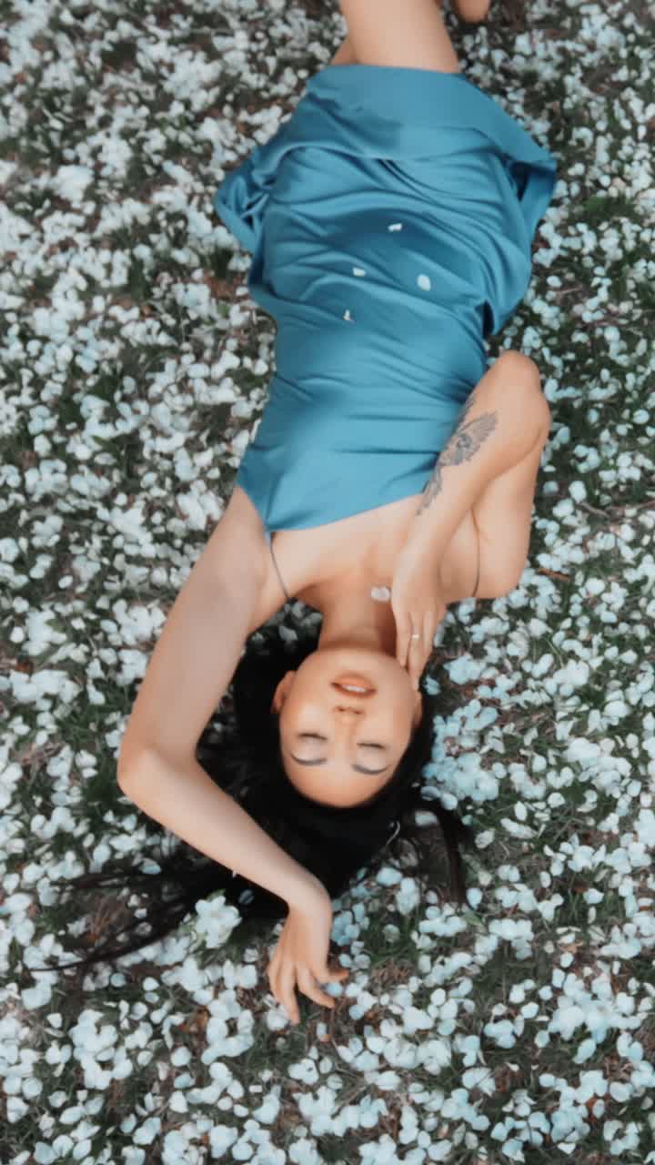 Woman in a blue silk dress lying on the grass covered in cherry blossoms