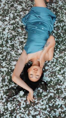woman in a blue silk dress lying on the grass covered in cherry blossoms