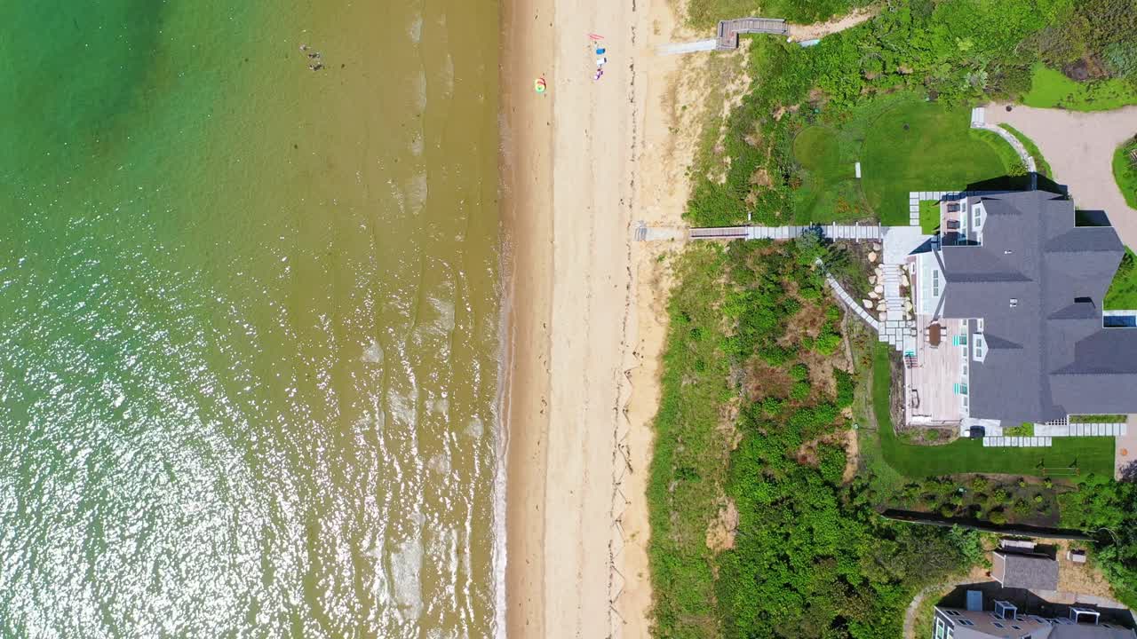 Overhead drone footage reveals beach houses tucked into greenery above sandy shoreline, with umbrellas dotting the beach and turquoise waters lapping against the coast