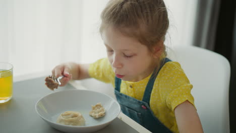 girl wearing yellow polka dot top sitting at dining table holding fork with food, snack dropping into plate, satisfied expression, bright cozy home environment with glass of juice nearby