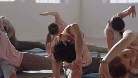 yoga class healthy mixed race woman practicing seated side bend pose with group of beautiful women sitting on exercise mat enjoying healthy lifestyle training in fitness studio at sunrise