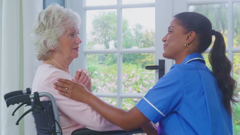 Female-Care-Worker-In-Uniform-Talking-With-Senior-Woman-Sitting-In-Wheelchair-In-Care-Home-Lounge