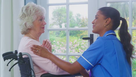 female care worker in uniform talking with senior woman sitting in wheelchair in care home lounge