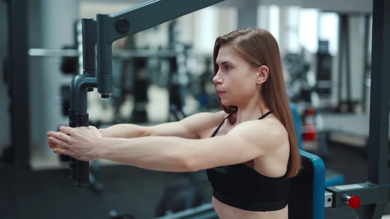 A beautiful girl with her strong arms performs an exercise on a simulator to strengthen the pectoral muscles in a fitness club. Close-up. Blurred Background