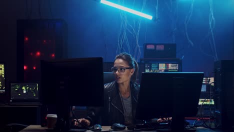 mujer joven con anteojos sentada y trabajando en la computadora en la habitación oscura con tecnologías, pirateando el sistema y lavando datos