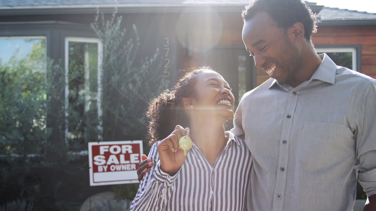 Portrait Of Couple Standing Outdoors In Front Of House With For Sale Sign In Garden Holding Keys