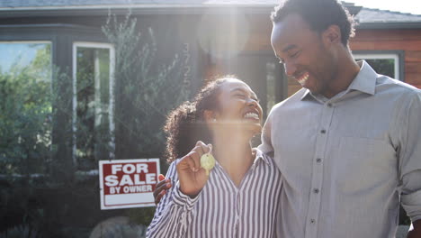 Portrait-Of-Couple-Standing-Outdoors-In-Front-Of-House-With-For-Sale-Sign-In-Garden-Holding-Keys