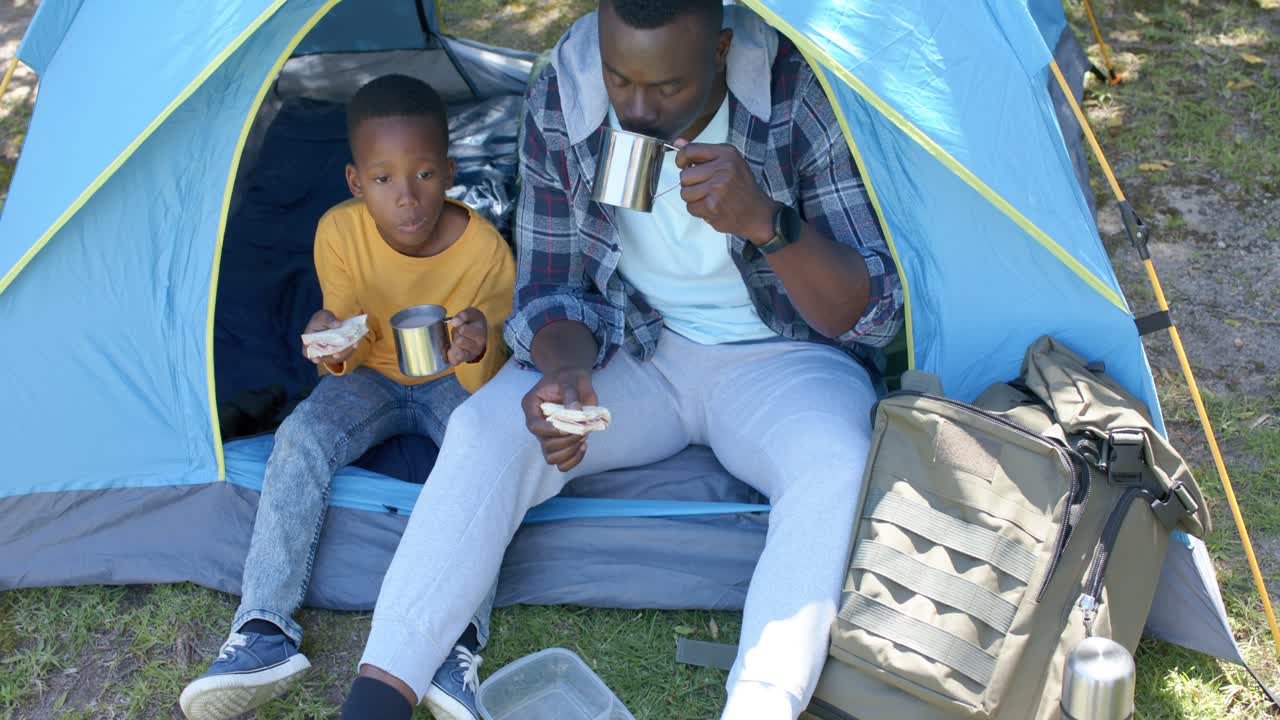 Afroamerikansk far og søn holder snackpause ved et blåt telt, manden tjekker en metalkedel.