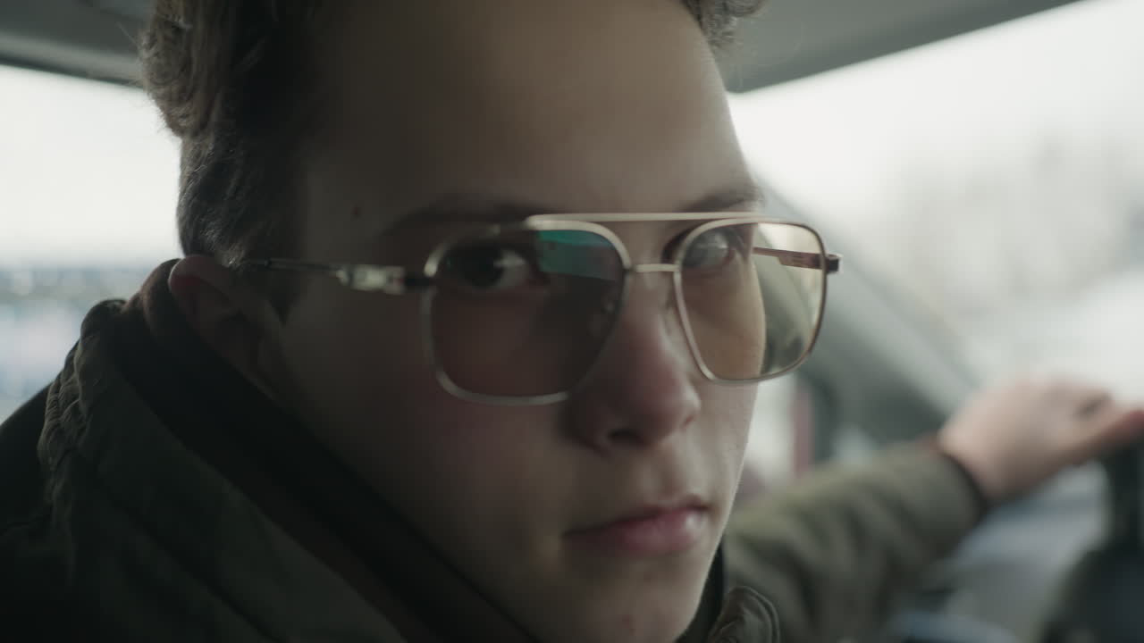 close up of teenager in glasses and winter jacket behind wheel turning head toward camera with serious expression as snowy cityscape appears blurred through car window in background