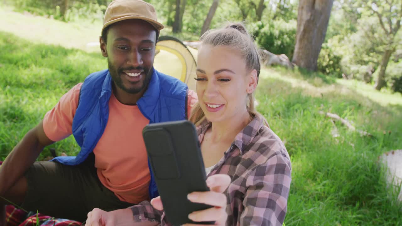 Happy diverse couple camping and taking selfie with smartphone in park, slow motion