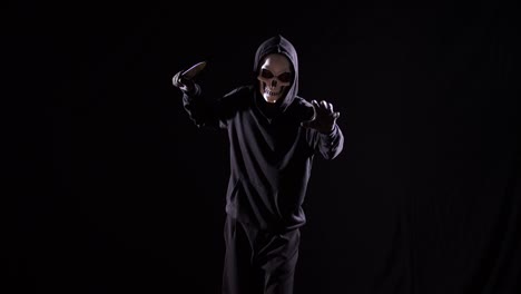 hombre aterrador en la sudadera con capucha con máscara de halloween sosteniendo un cuchillo y haciendo un gesto aterrador mientras camina sobre el fondo negro