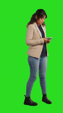 a young woman in casual wear stands in front of a green screen while looking down at her phone and smiling.