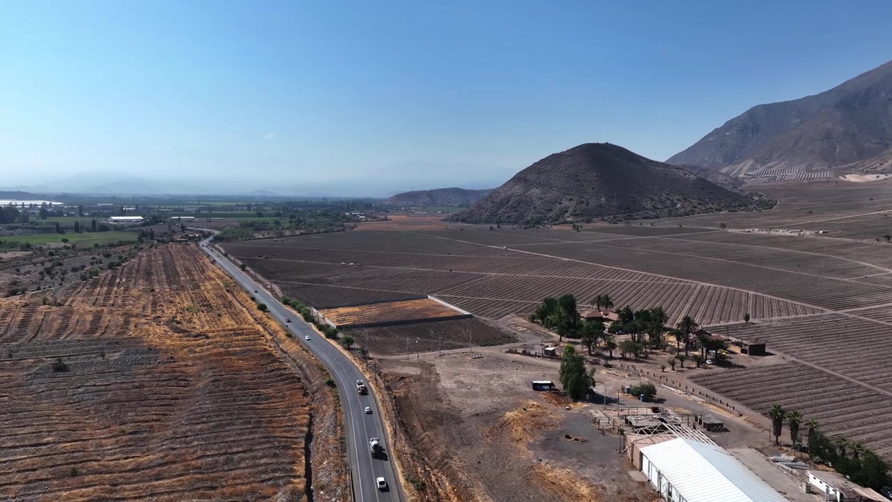 Vista aerea panoramica su una valle agricola arida con campi mietuti, strada tortuosa, complesso agricolo rurale e collina vulcanica, sullo sfondo una drammatica catena montuosa andina sotto un cielo blu.