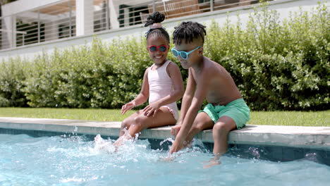 Hermana-Y-Hermano-Afroamericanos-Disfrutan-De-La-Diversión-Junto-A-La-Piscina-En-Casa,-Salpicando-Agua-Con-Los-Pies