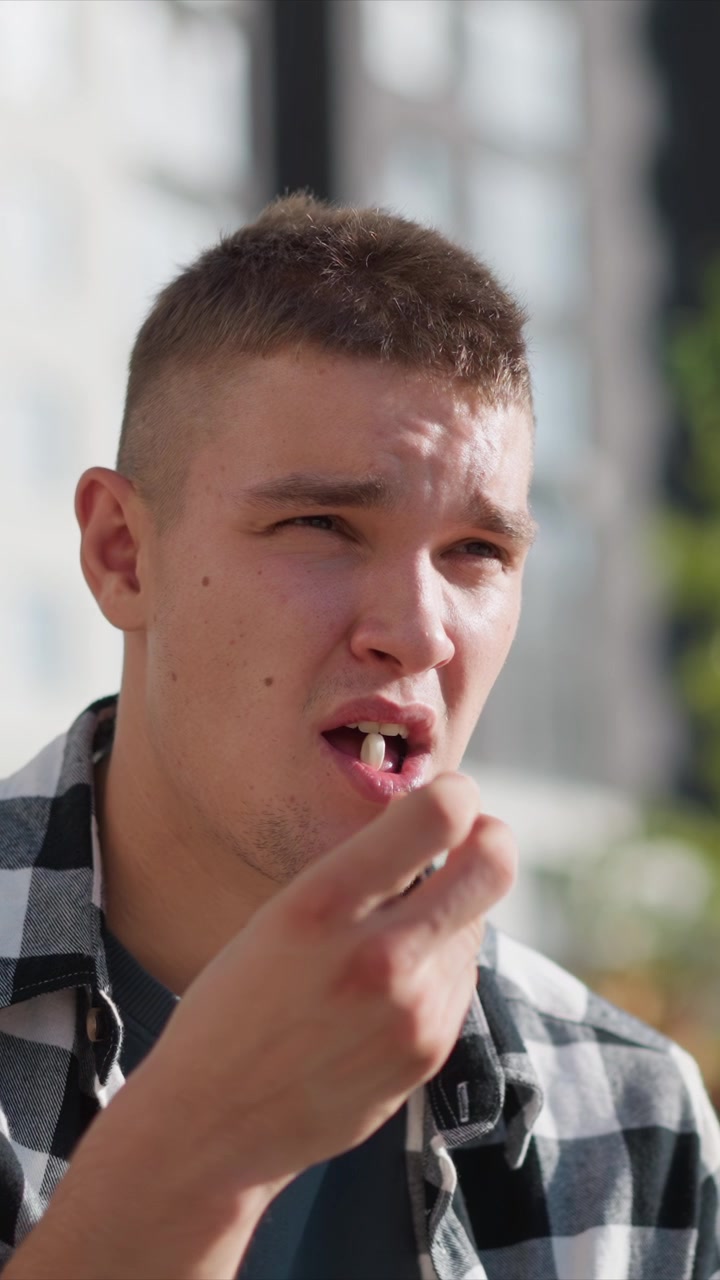 Close-up de um jovem falando ao ar livre com leves gestos de mão, vestindo camisa xadrez e camiseta escura. A luz natural suave realça suas características faciais contra um fundo urbano.