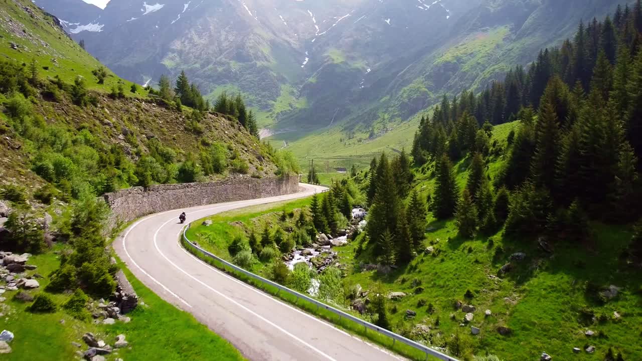 Drone tracks biker riding through Transfăgărășan road in Carpathian Mountains of Romania, surrounded by lush greenery and alpine landscape