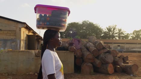 young woman with some load on her head walking towards the right at a sawmill compound may be peddling some drinks to the commnuity in a kumasi, ghana