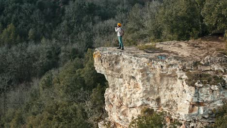 aerial view of a woman on the edge of a cliff taking landscape photos by a river, she is wearing an orange cap