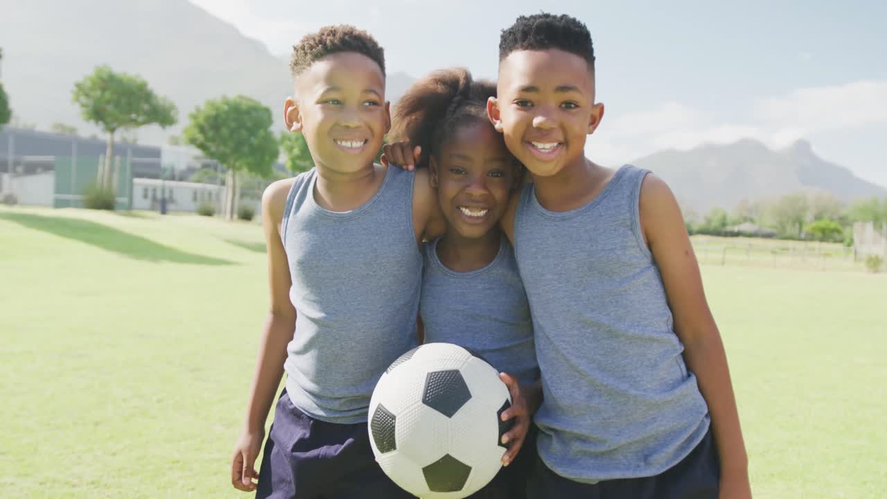 retrato de video de tres niños afroamericanos felices sosteniendo un fútbol en el campo de la escuela