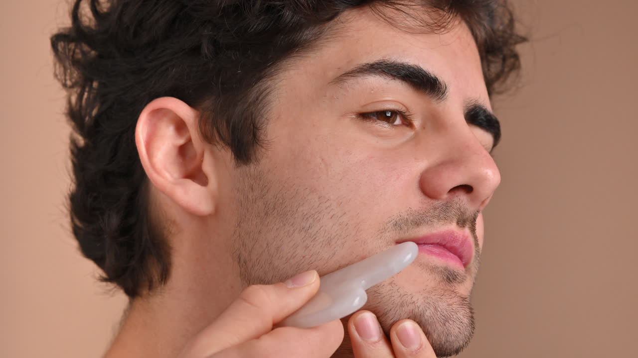 A young caucasian man with stubble beard is doing a facial massage using a Gua Sha