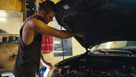 a male master motorist mechanic with a short haircut in a gray t-shirt checks the hood of the car and the oil there in his garage workshop. car hood repair in my workshop