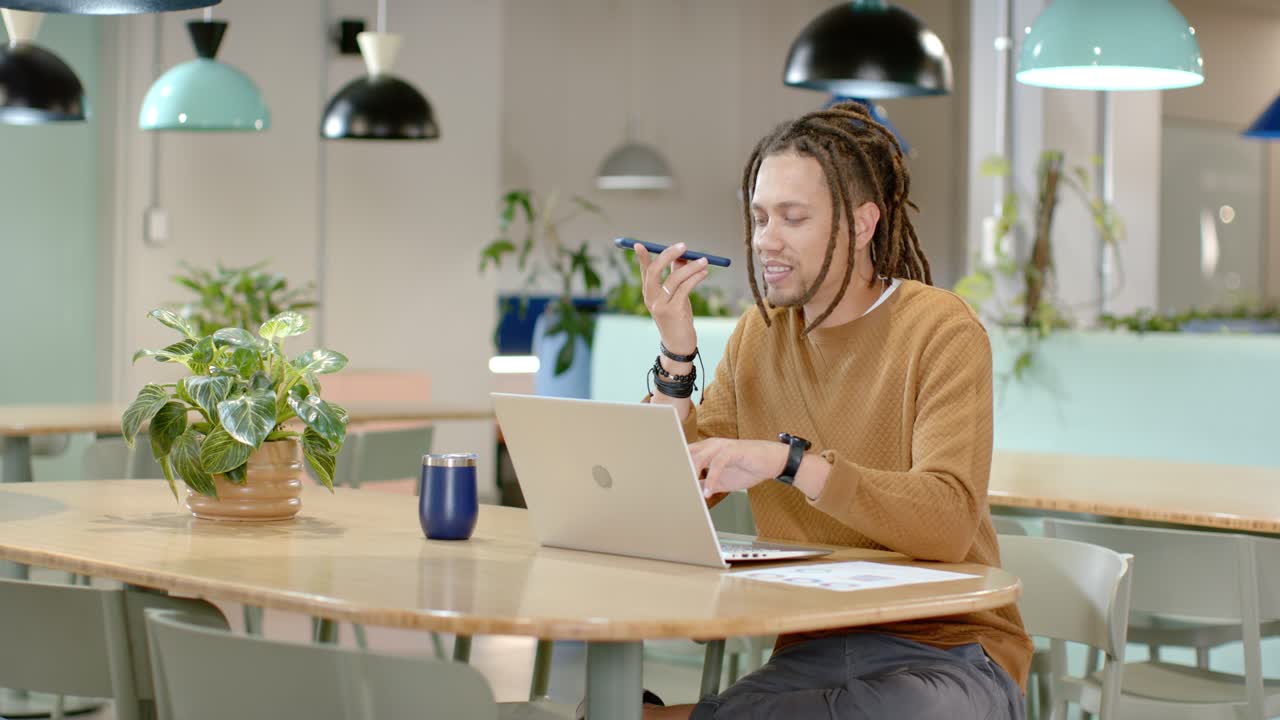 African American man sitting at coworking table lifting phone to mouth, speaking, typing on laptop