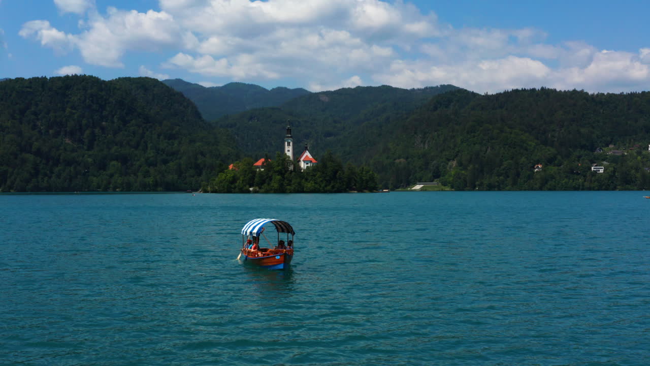 Turisti in crociera sul Lago di Bled a bordo di una barca Pletna, con vista sulla Chiesa di Santa Maria Assunta e sulla catena montuosa in Slovenia