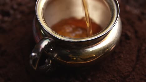 Swirling-coffee-pouring-into-ornate-silver-cup-rotating-on-shallow-focus-dark-ground-aromatic-coffee-beans-close-up