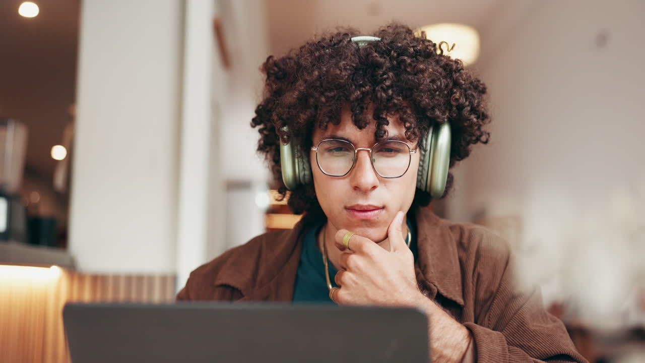 Man with curly hair wearing headphones and glasses working on a laptop