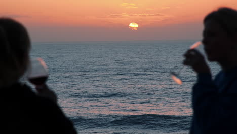 silhouette of two girls drinking a red wine in front of beautiful ocean and golden sunset in background