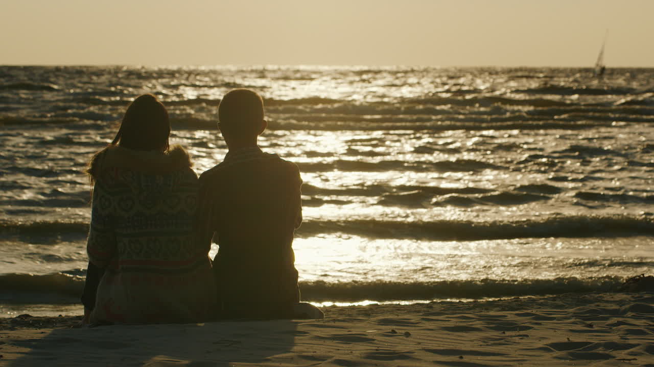 일몰을 바라보며 해변에 앉아 사랑에 빠진 젊은 부부와 윈드서핑을 타는 사람