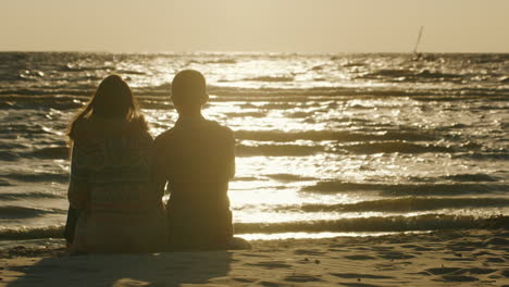 Junges-Verliebtes-Paar-Sitzt-Am-Strand-Und-Beobachtet-Den-Sonnenuntergang-Und-Die-Person,-Die-Auf-Windsurfen-Reitet
