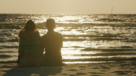 Young-Couple-In-Love-Sitting-On-The-Beach-Watching-The-Sunset-And-The-Person-Who-Rides-On-Windsurfin