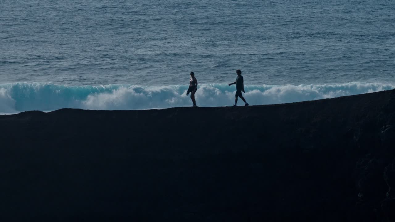 A young couple walks hand in hand along a rugged volcanic cliff overlooking the Atlantic Ocean in Lanzarote, Canary Islands, Spain.