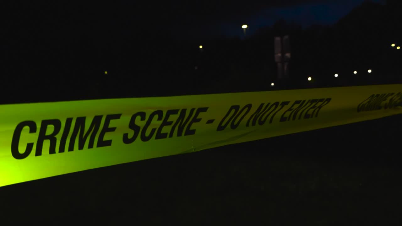 Close up or closeup of a yellow colored Crime Scene Do Not Enter police tape ribbon moving in the wind in front of a dark background with bokeh street light during evening or night time, well lit tape