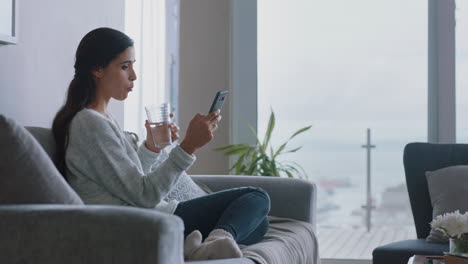 Schöne-Junge-Frau-Nutzt-Ihr-Smartphone,-Entspannt-Sich-Zu-Hause-Auf-Der-Couch-Und-Surft-Online,-Schaut-Sich-Unterhaltung-An,-Trinkt-Wasser-Und-Genießt-Einen-Komfortablen-Lebensstil