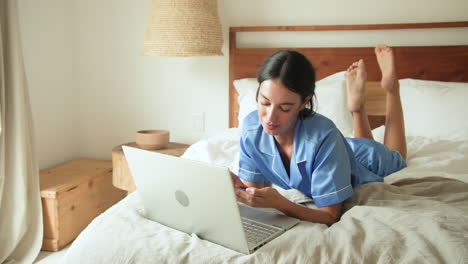 at home, woman in pajamas using laptop on bed, looking focused and relaxed