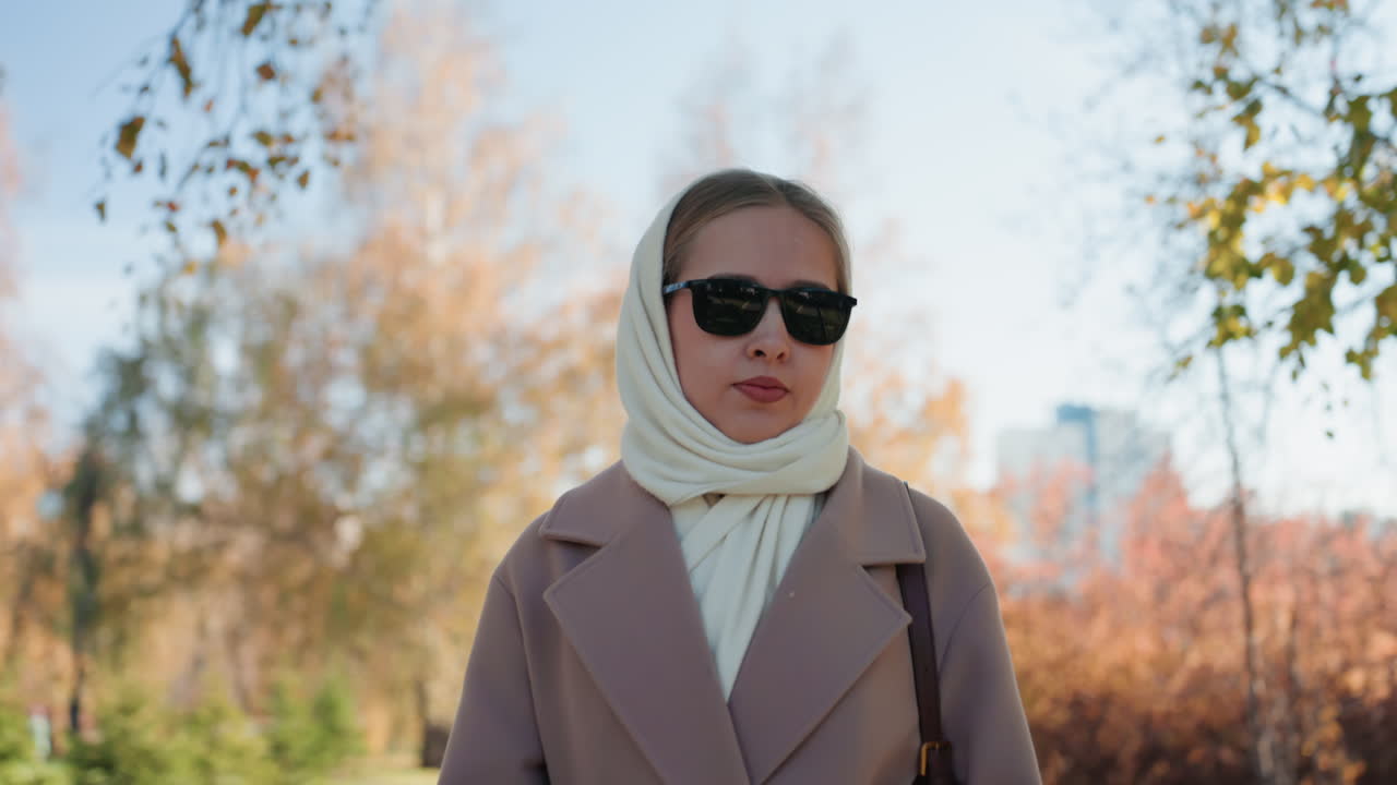 Donna caucasica con occhiali da sole passeggia con sicurezza in un parco autunnale, cappotto beige e sciarpa color crema, postura chic, luce solare attraverso il fogliame colorato, energia da fashion blogger, scena urbana rilassata.