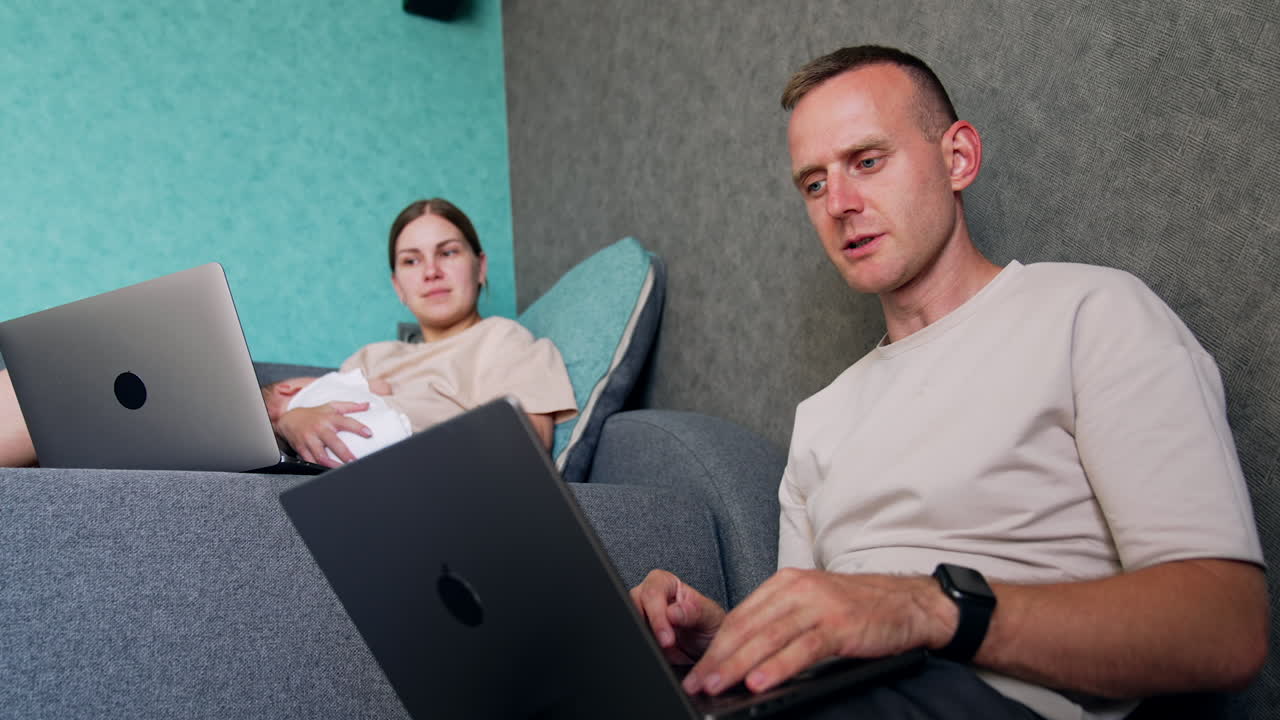 Young couple having a newborn baby working at home. Two parents work on laptops and discuss work. Low angle view.