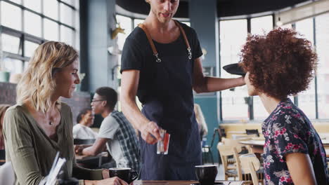 Two-Female-Friends-Sitting-At-Table-In-Coffee-Shop-Being-Served-By-Waiter