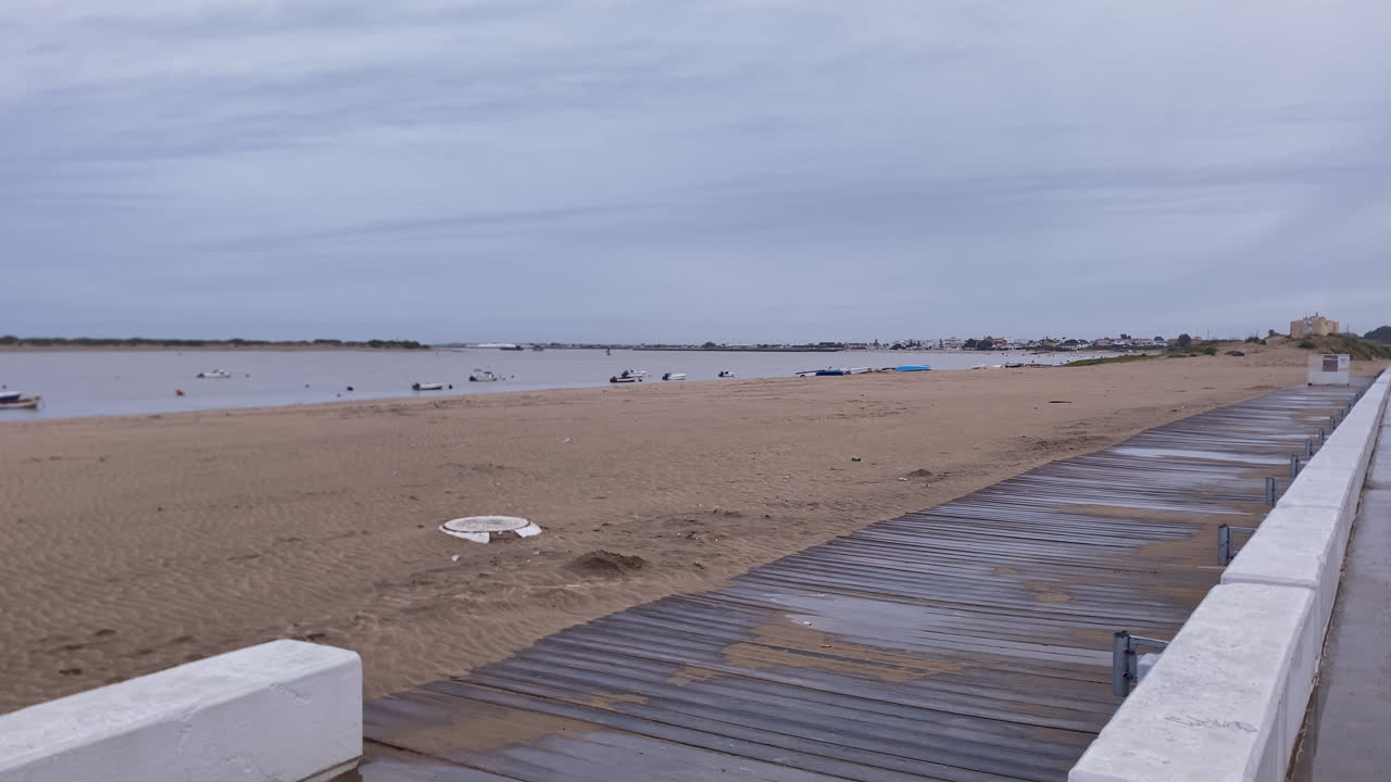 Sandy beach alongside the Guadalquivir river mouth with a boardwalk and anchored boats under a cloudy sky