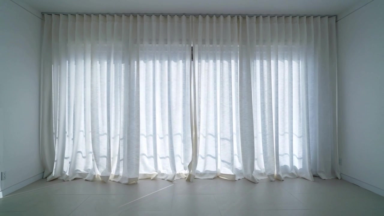 Cortinas altas y transparentes ondeando, respondiendo a la luz solar cambiante detrás del cristal en una habitación blanca vacía.
