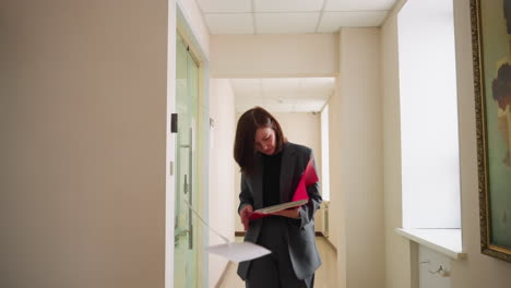 businesswoman walking down office corridor, reviewing documents in folder, professional attire, modern office, focused on task, corporate environment, businesswoman reading papers