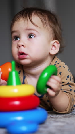 lovely boy lies on carpet indoors with a toy in front of him. cute kid gathering the parts of bright toy pyramid. blurred backdrop. vertical video