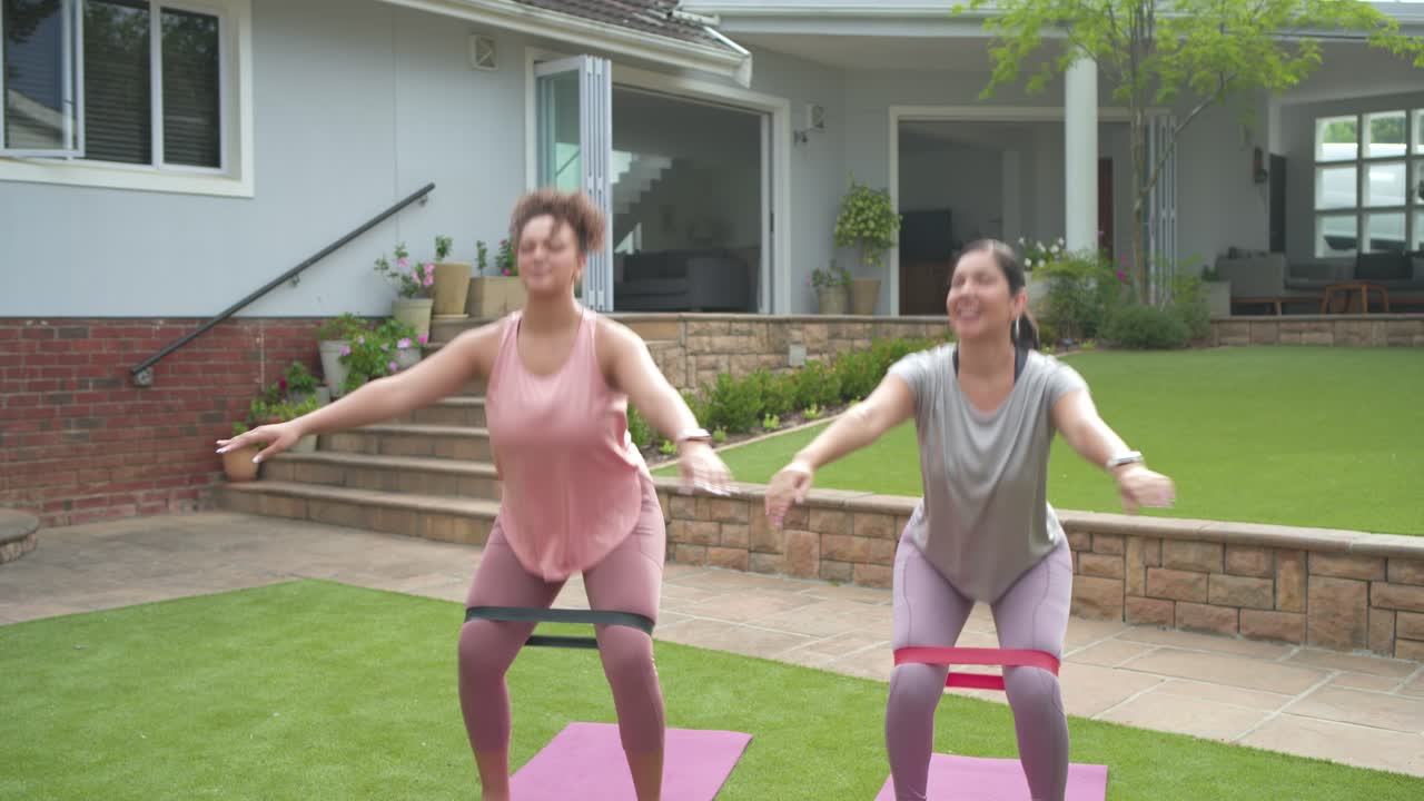 Diversas amigas comienzan sentadillas tras una sonrisa compartida, haciendo sentadillas con banda en el césped para mantenerse en forma.