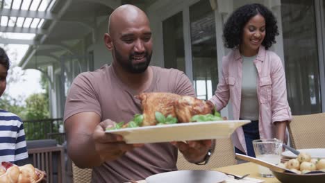 happy biracial parents and son serving meal at dinner table in garden, slow motion