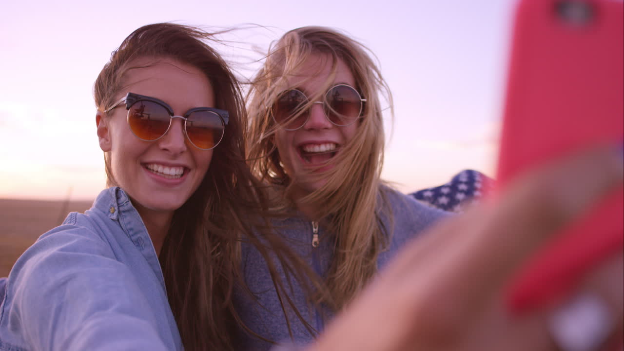 Belle amiche che si scattano selfie durante un viaggio on the road al tramonto con un'auto d'epoca