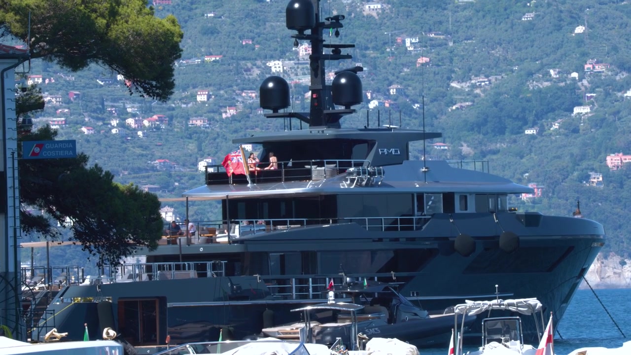 Un grande superyacht grigio scuro è ancorato in un soleggiato porto turistico italiano con colline lussureggianti.